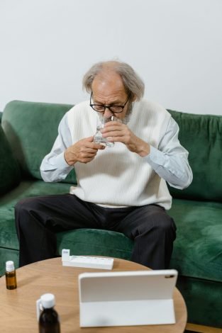 Senior man sitting on a green couch drinking water and taking medication, emphasizing health and wellness.